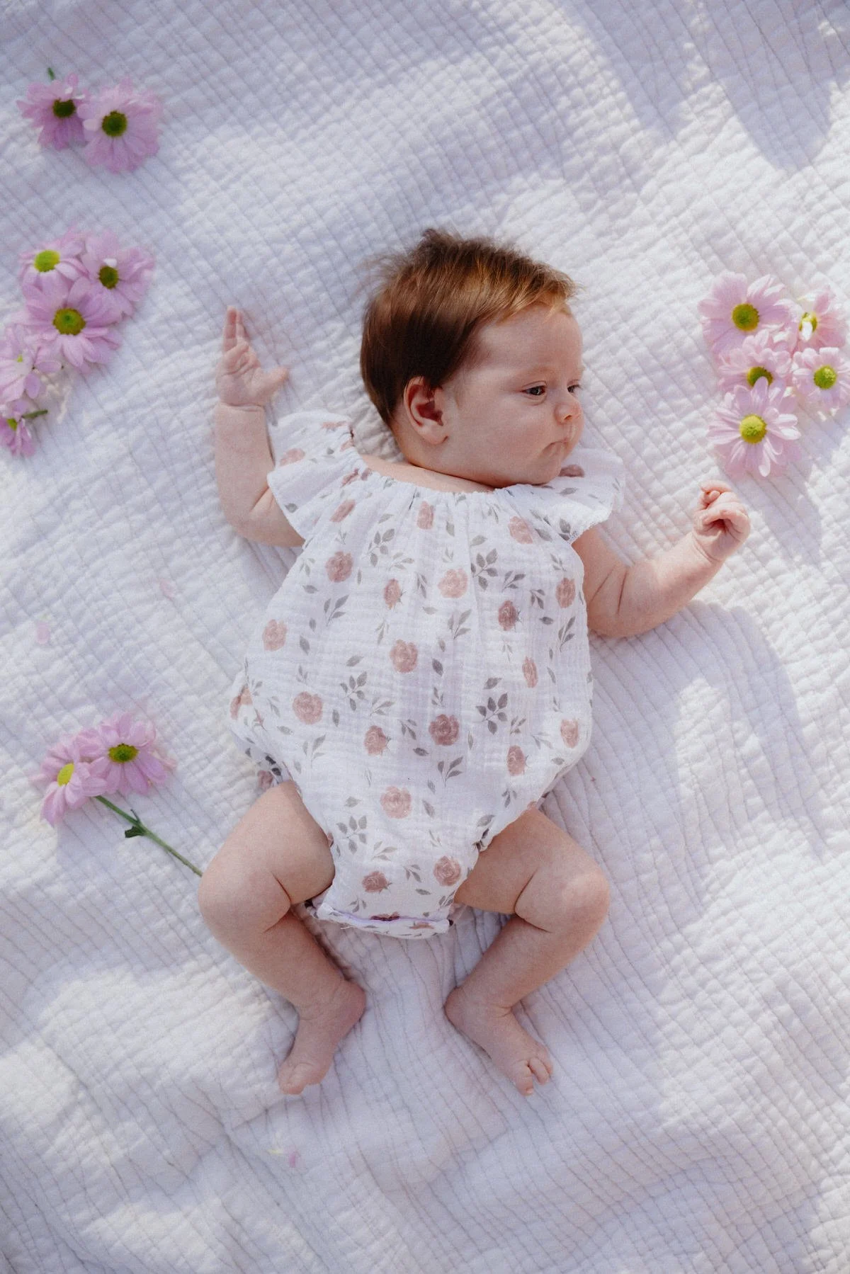 A baby with red hair lying on a white quilted blanket, surrounded by pink daisies, wearing a white dress with pink and gray floral patterns.
