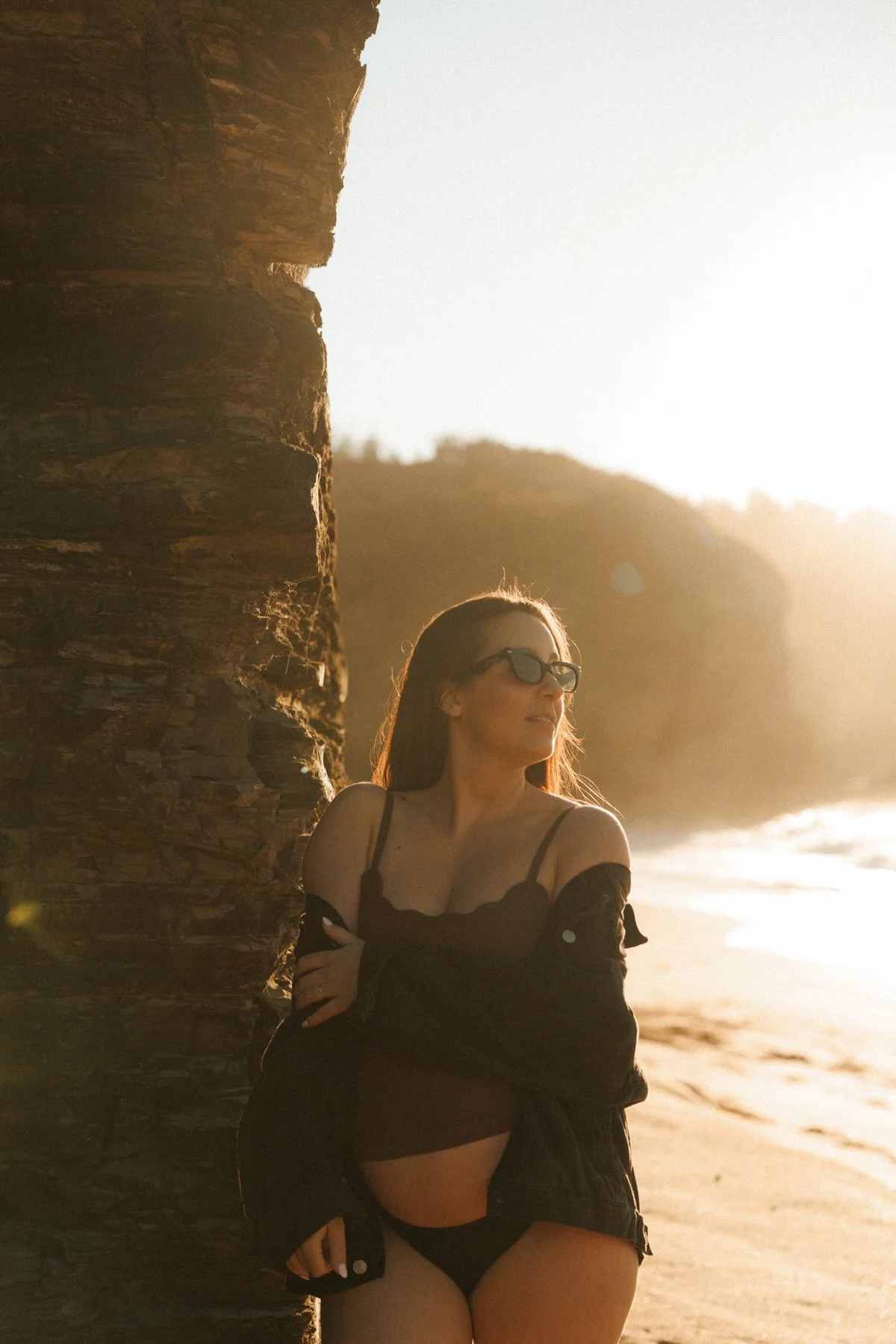 A woman with long hair and sunglasses stands against a large rock formation at the beach during sunset, wearing a black off-shoulder top and shorts, with the ocean and a hilly landscape in the background.