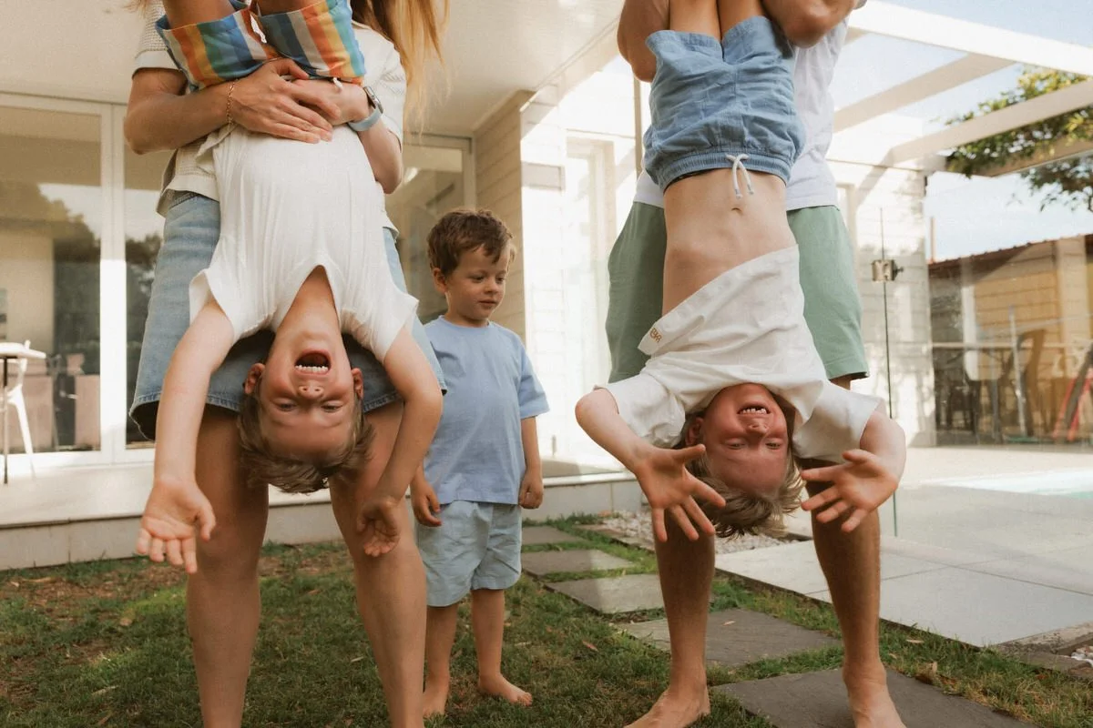 Two children are hanging upside down from their parents' legs, smiling and making funny faces. A young boy in a blue shirt is standing nearby, watching and smiling, while an adult is holding one of the children. They are outside in a backyard with a 
