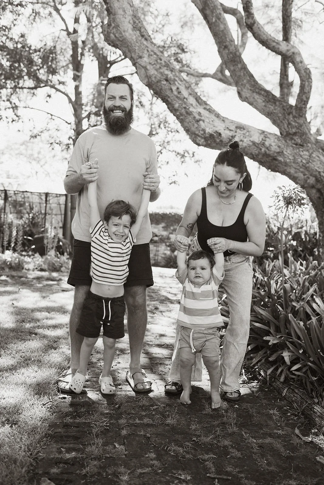 A happy family of four outdoors, with a man, woman, and two young children, standing on a dirt path near a large tree, smiling and holding hands, in a garden or park setting.
