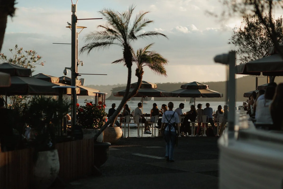 People dining at outdoor restaurant by the water with palm trees, umbrellas, and a scenic view of a lake or ocean during sunset.