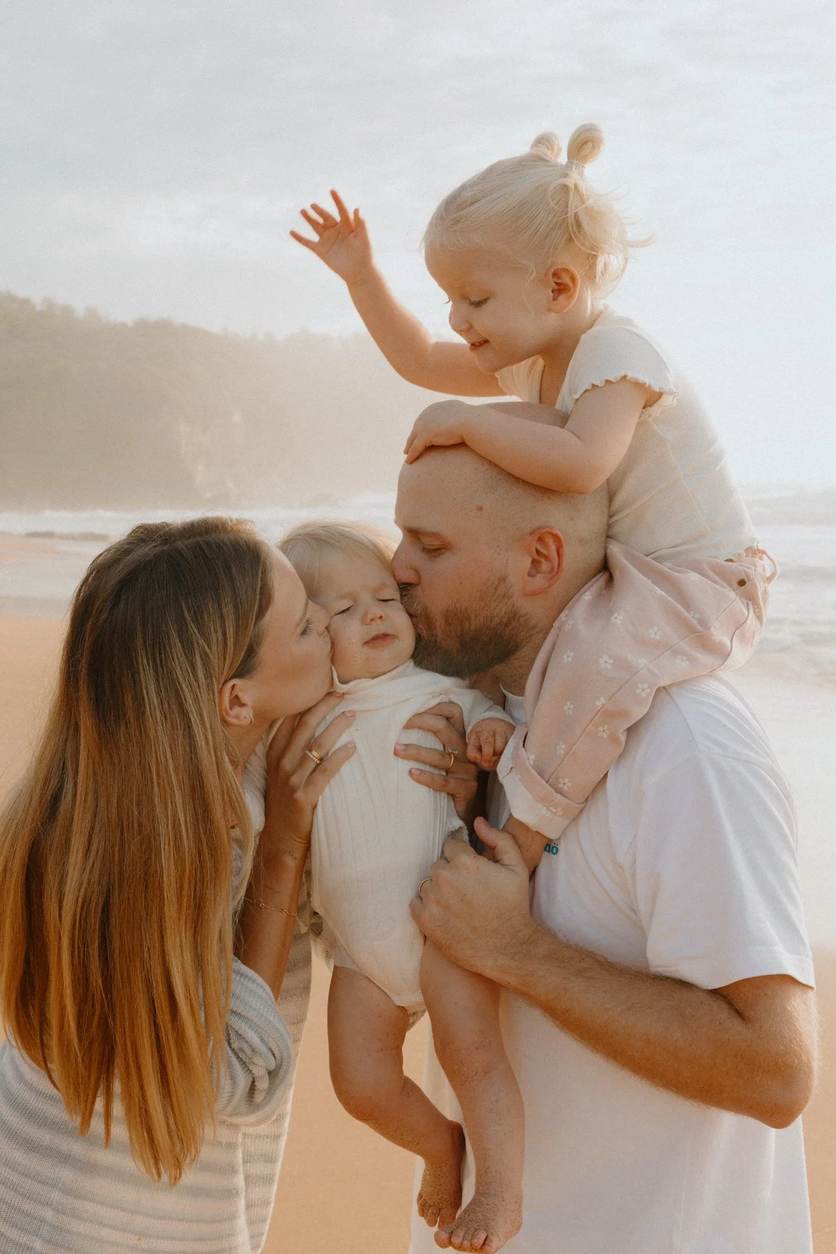 A family of five at the beach, with the parents kissing one of their children, while the two young girls smile and wave.