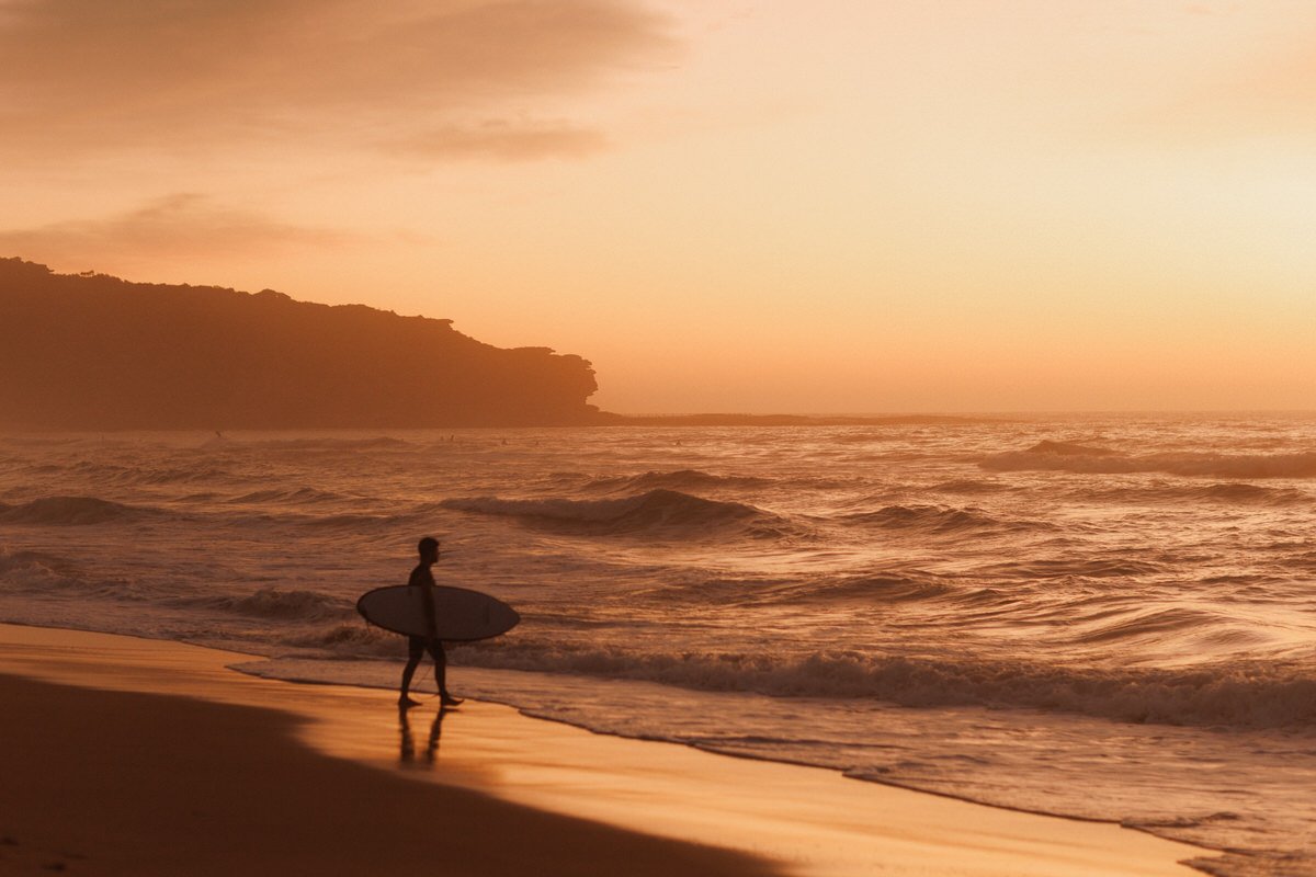 A person holding a surfboard walking along the sandy beach during sunset with the ocean waves in the background.