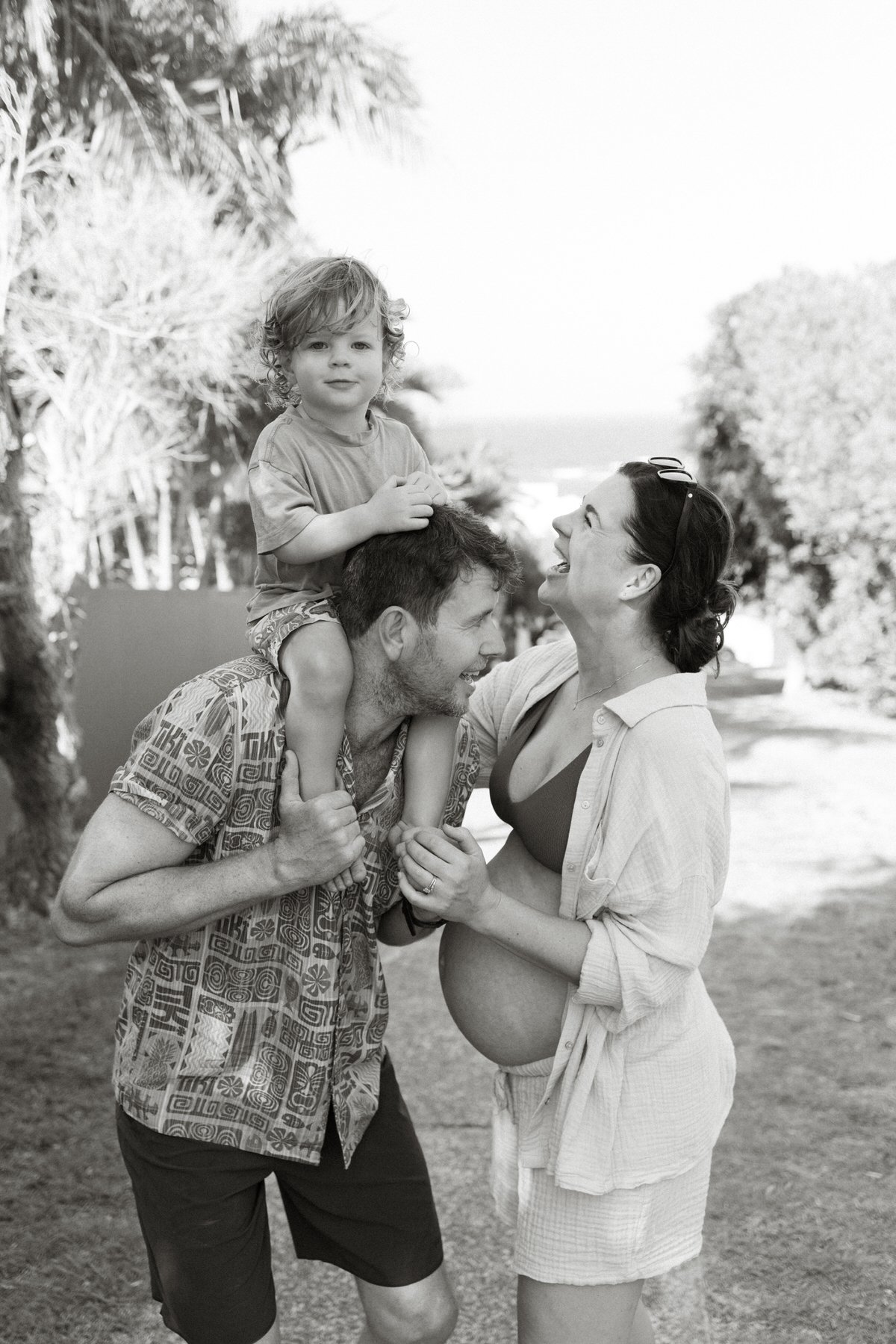 A family enjoying a moment outdoors with the father carrying a young boy on his shoulders and the mother, who is pregnant, smiling at them.