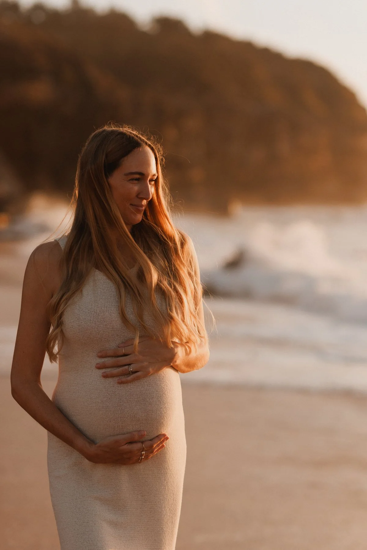 A pregnant woman with long, wavy hair standing on a beach during sunset, smiling gently and holding her belly with both hands.