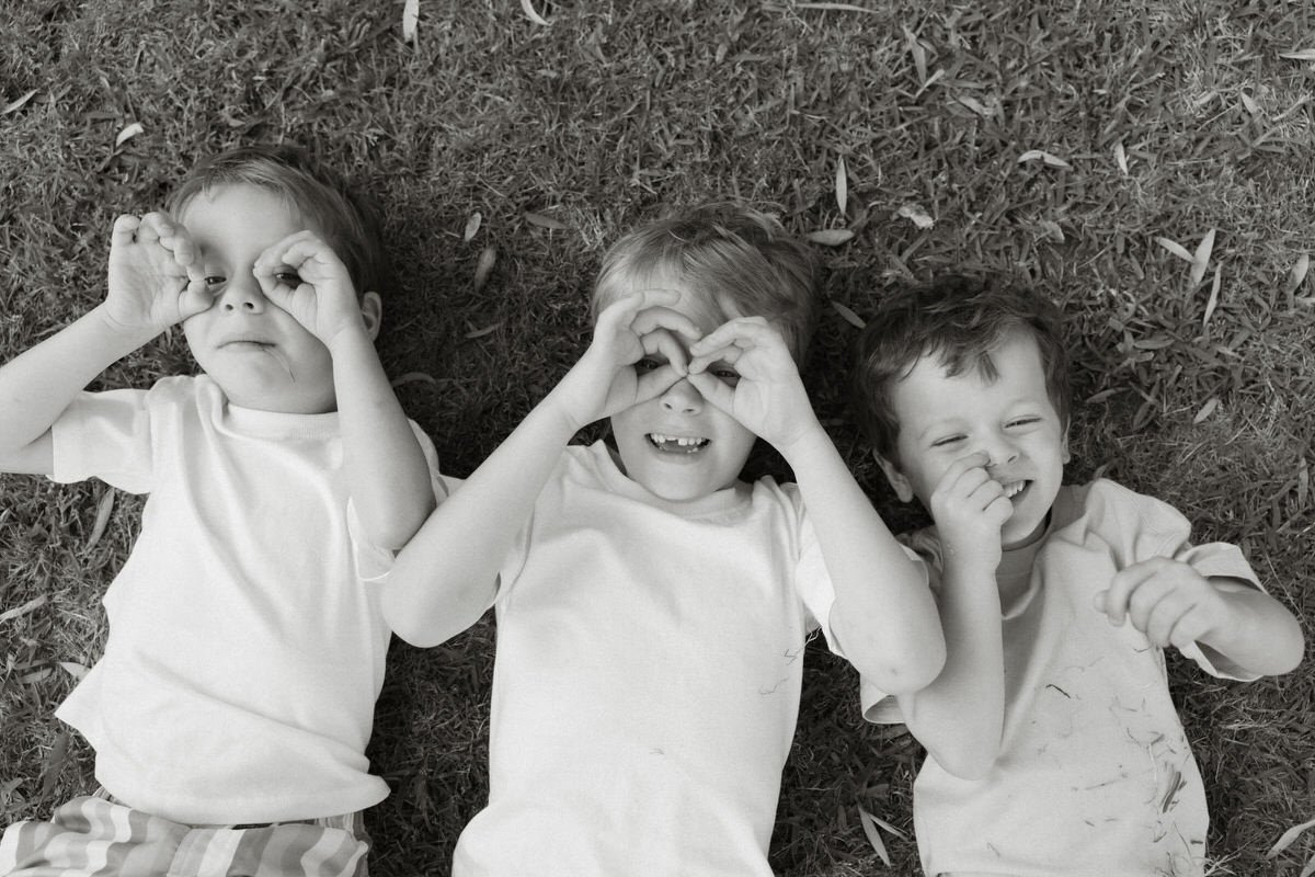 Three young boys lying on grass, making silly faces and playful gestures.