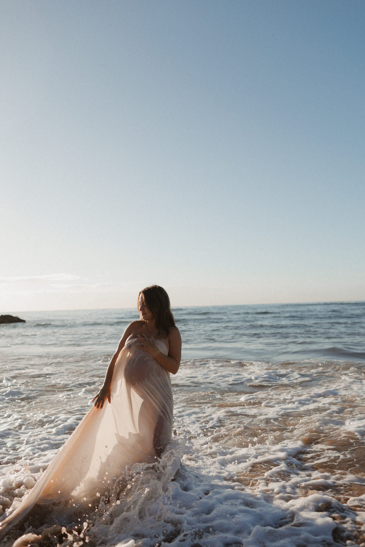 A woman in a flowing dress stands in shallow ocean water with waves around her, under a clear sky.
