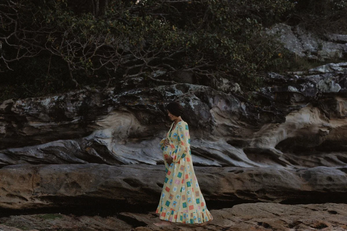 A woman wearing a long, colorful patterned dress standing on rocky terrain near a large rock formation with trees and bushes overhead.