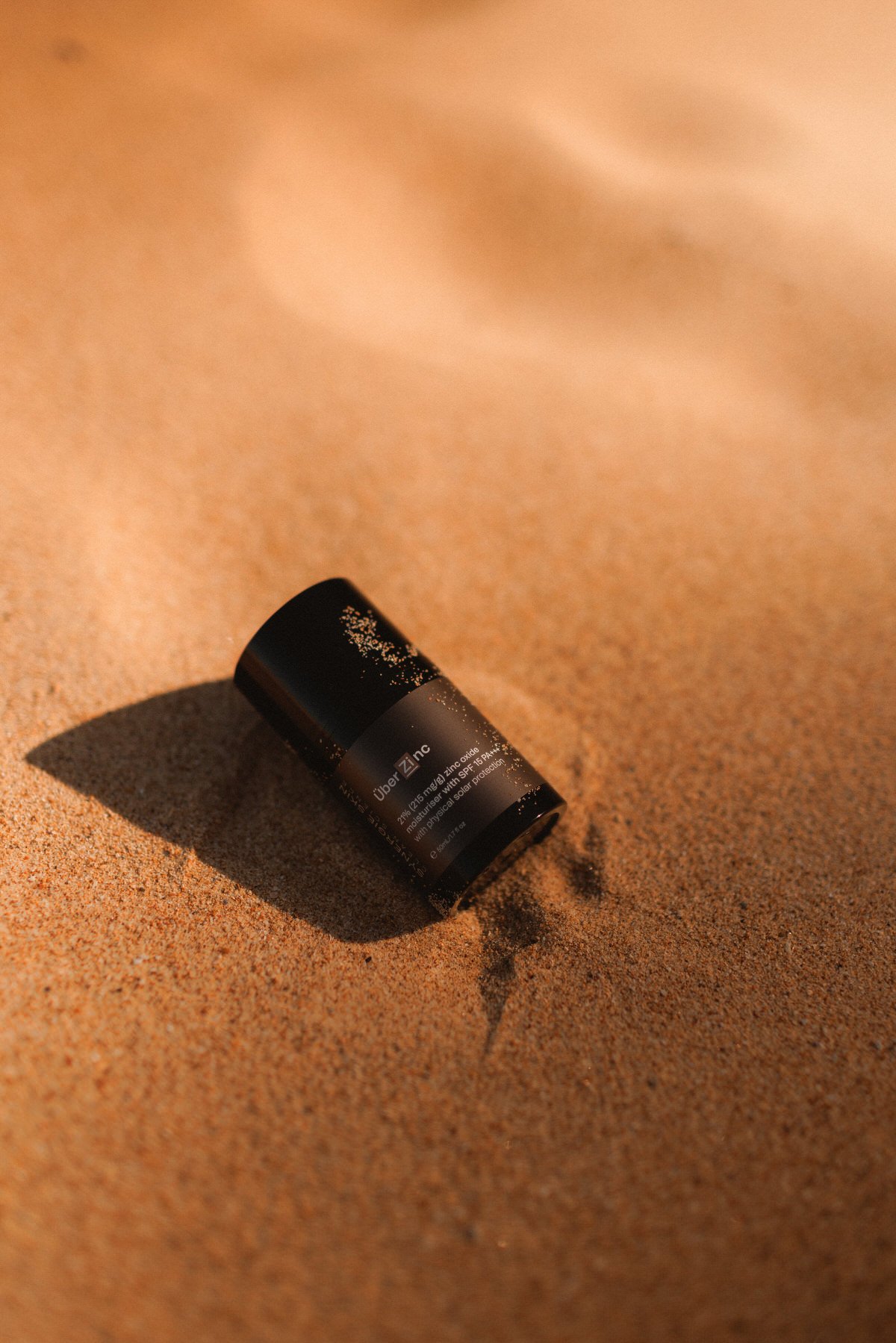 A small black container of Uber Zinc on sandy terrain, casting a shadow.