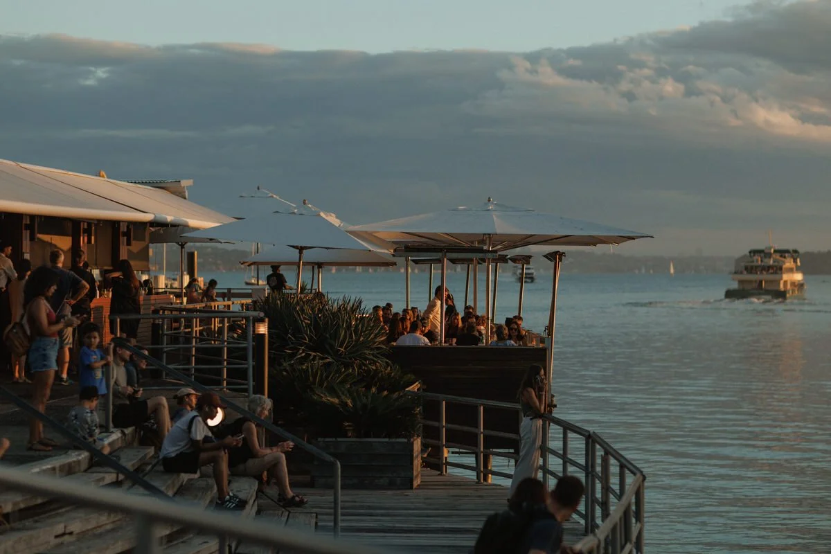 People sitting and standing on a lakeside deck, some under umbrellas, enjoying the sunset with boats on the water in the background.
