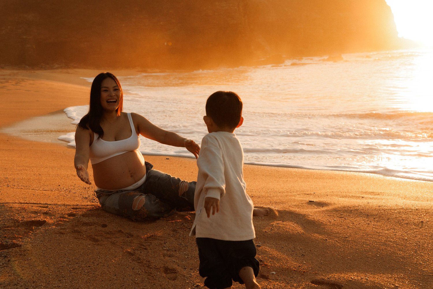 Pregnant mother sitting on the sand with open arms as her toddler runs towards her in Sydney winter morning light.