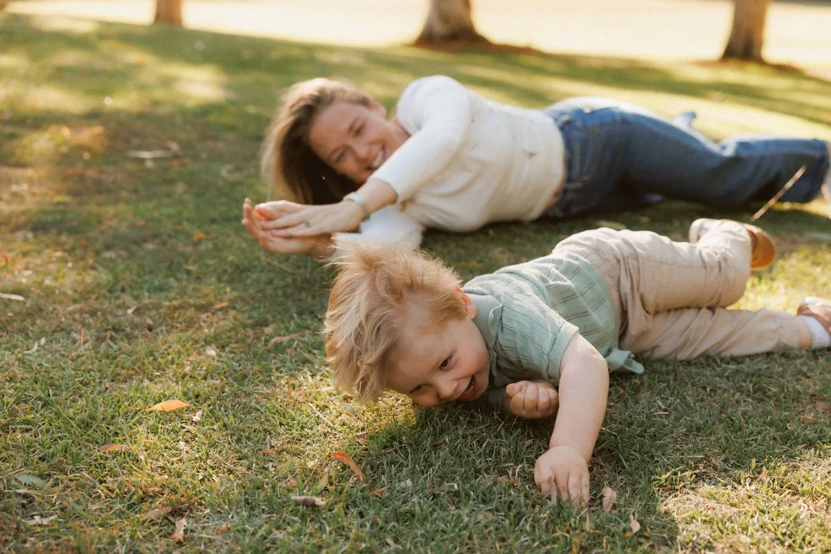 A woman lying on the grass smiling at a young boy crawling on the grass outdoors on a sunny day.