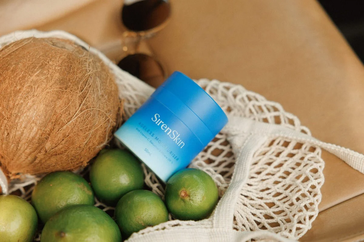 A blue container of SirenSkin moisturizer resting inside a white mesh bag surrounded by green limes and a brown coconut on a leather surface.