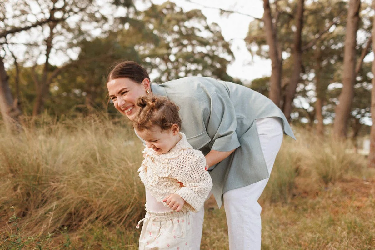 A woman and a young girl are outdoors in a grassy area with trees in the background. The woman is smiling and bent over, holding the girl by the waist. The girl appears to be looking down and is also smiling, wearing a cozy sweater and pants.