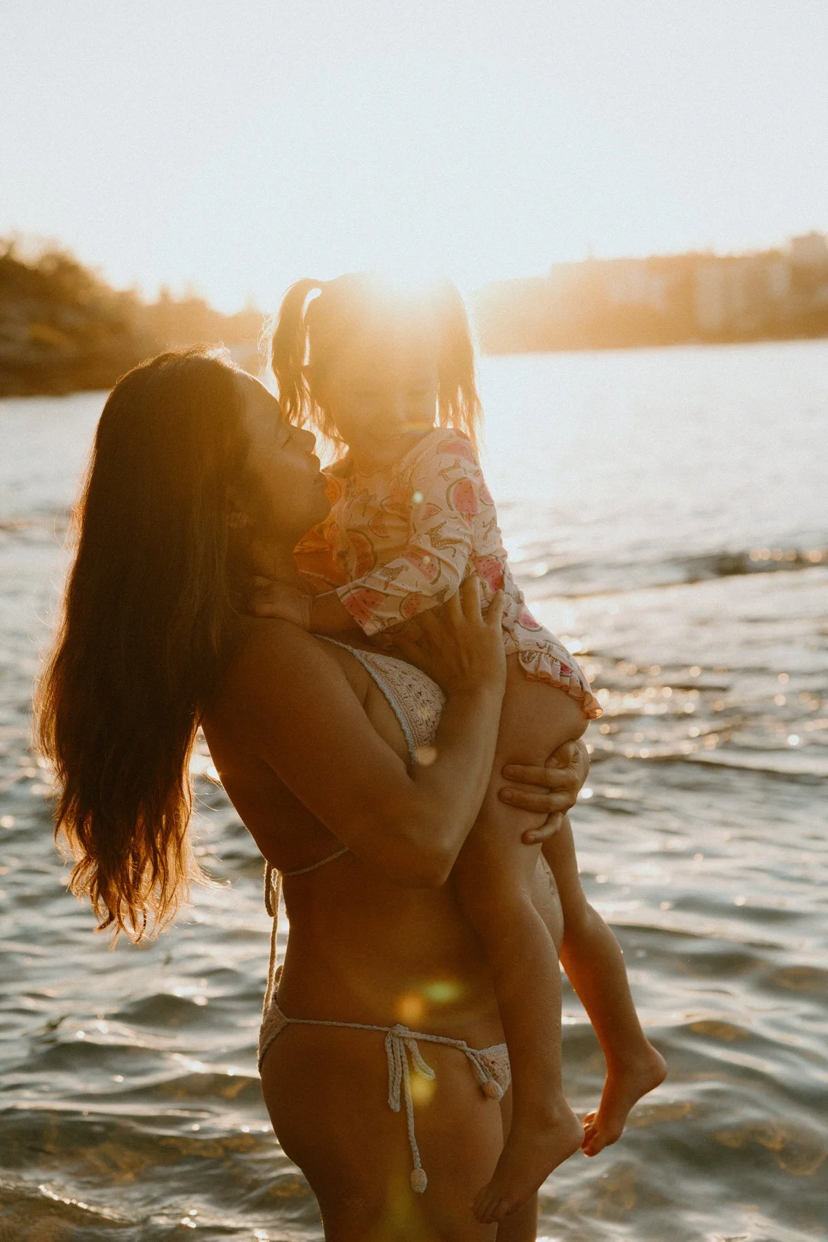 A woman holding a young girl in her arms at the beach during sunset. The sun is behind them, creating a bright glow over the water.