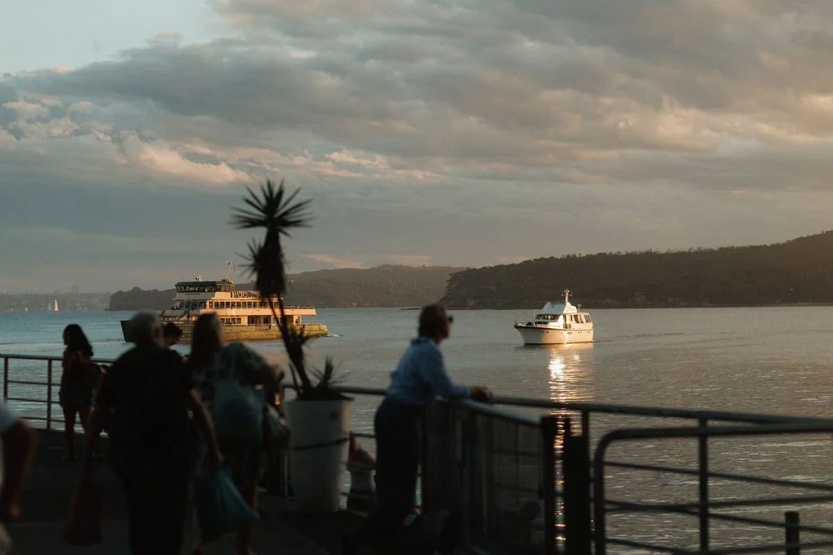 People standing on a waterfront promenade watching boats on the water at sunset, with hills and cloudy sky in the background.