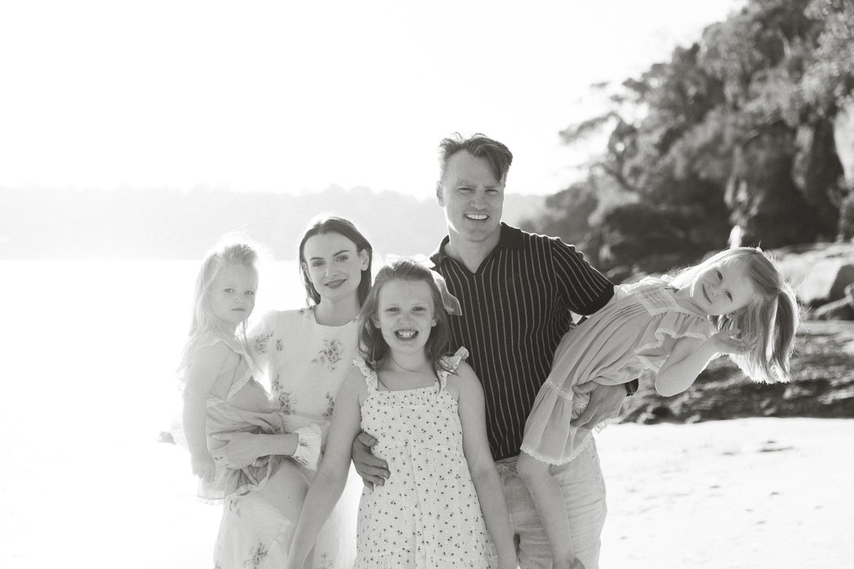 A family of five smiling at the beach, with rocky and tree-covered shoreline in the background.