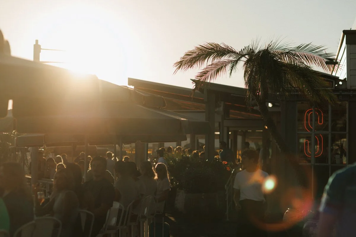 People are gathered outside a restaurant or cafe at sunset with a palm tree and neon signs in the background.