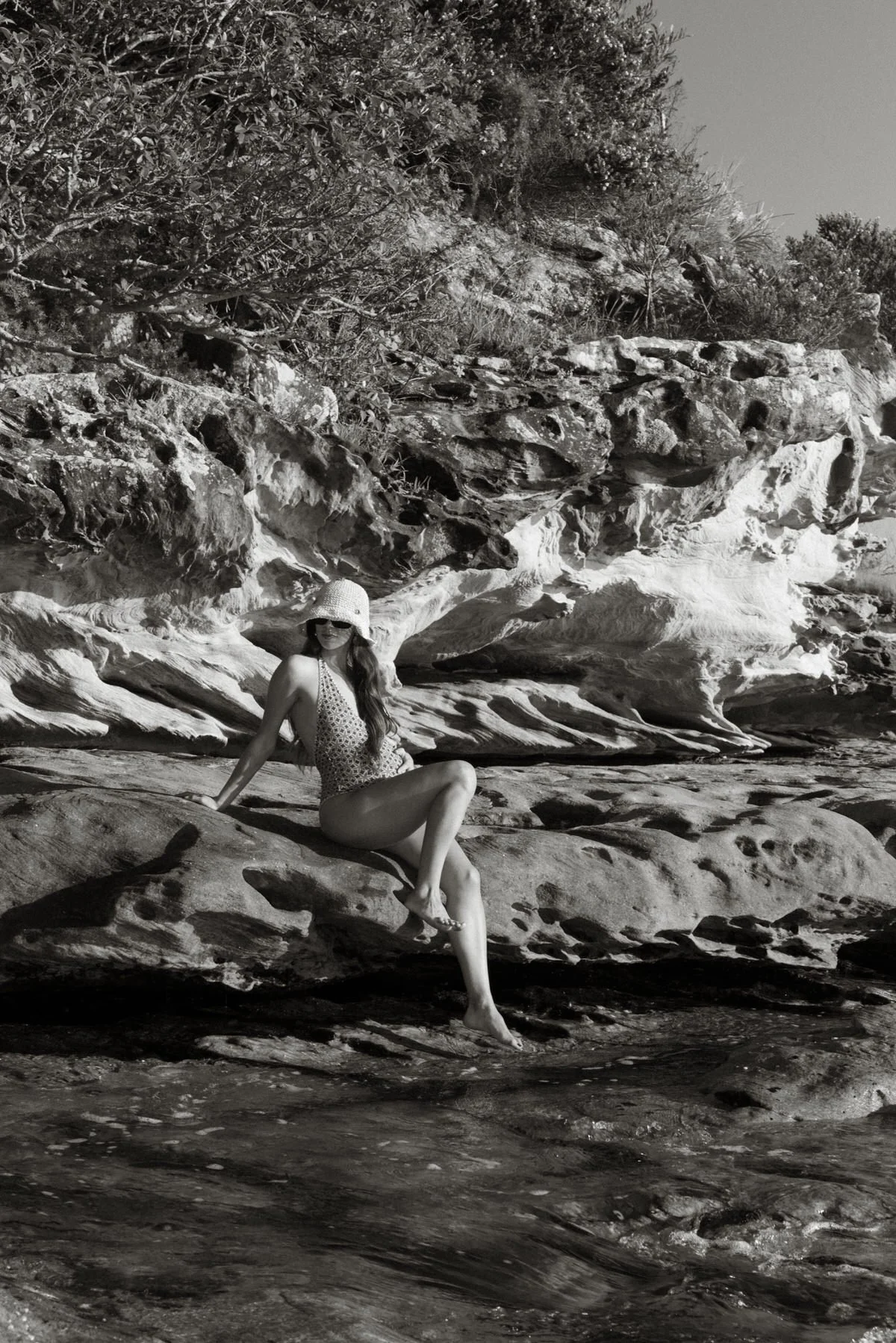 A woman in a swimsuit and sun hat sitting on rocks at the beach with a rocky cliff and bushes behind her.