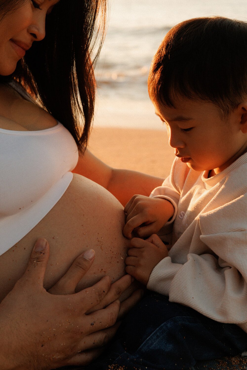 Close-up of a toddler gently touching their mother’s baby bump with both hands.