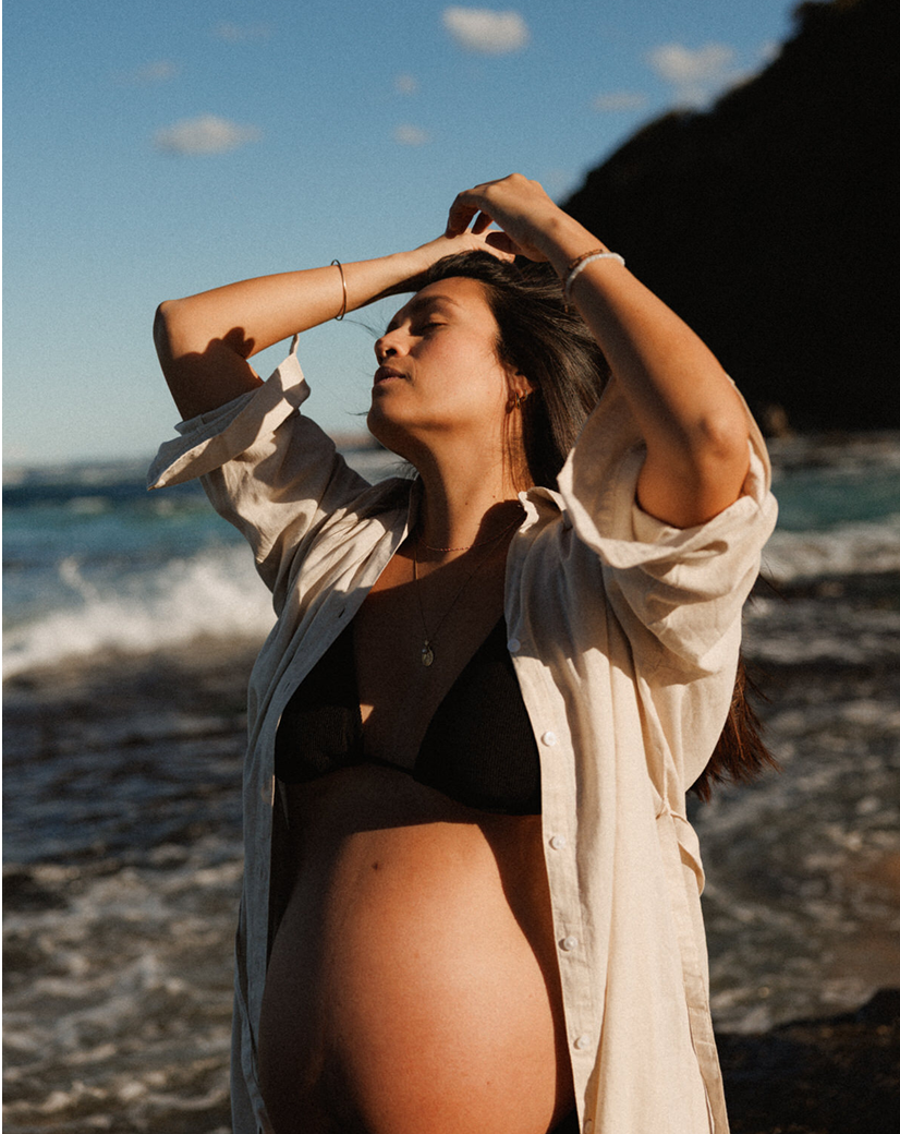 A woman standing on a beach with her eyes closed and her arms raised above her head, enjoying the sunlight with ocean waves in the background.