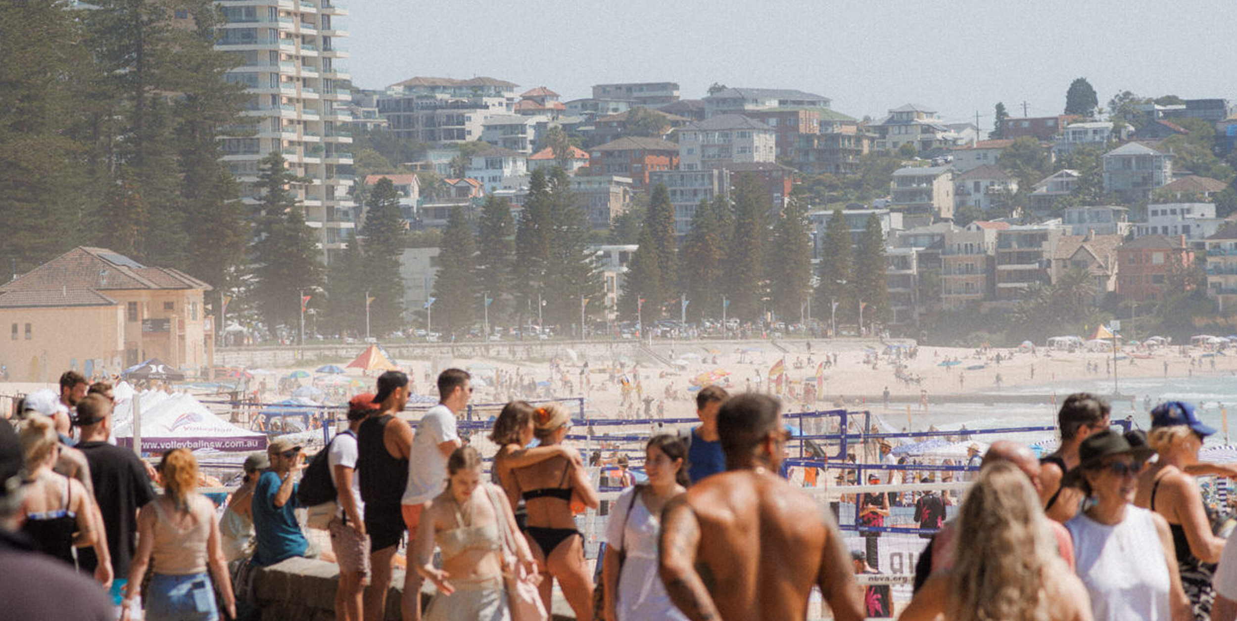 Crowd of people walking along a beach with a cityscape and hillside residential buildings in the background.