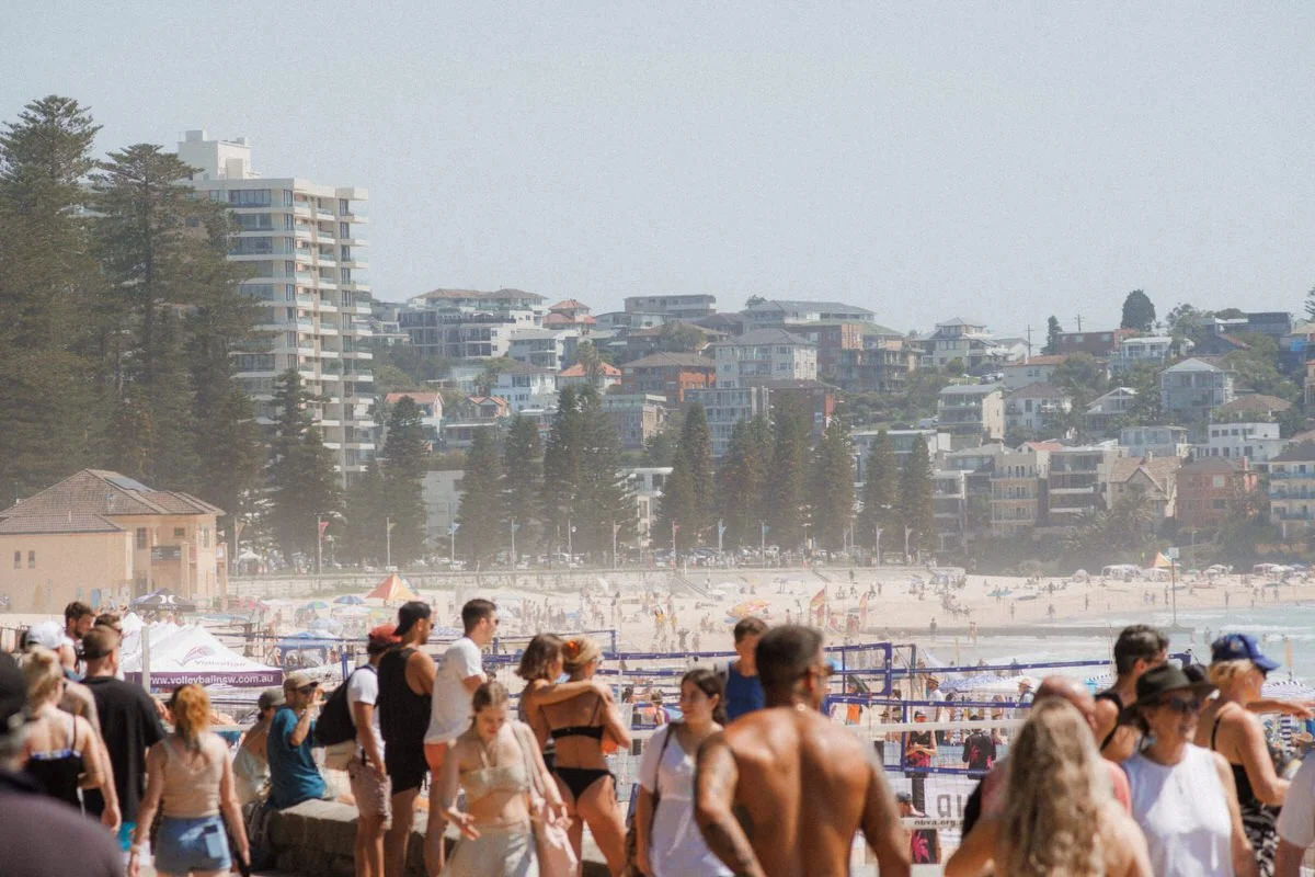 Crowd of people at a beach near high-rise buildings and trees with a cloudy sky.