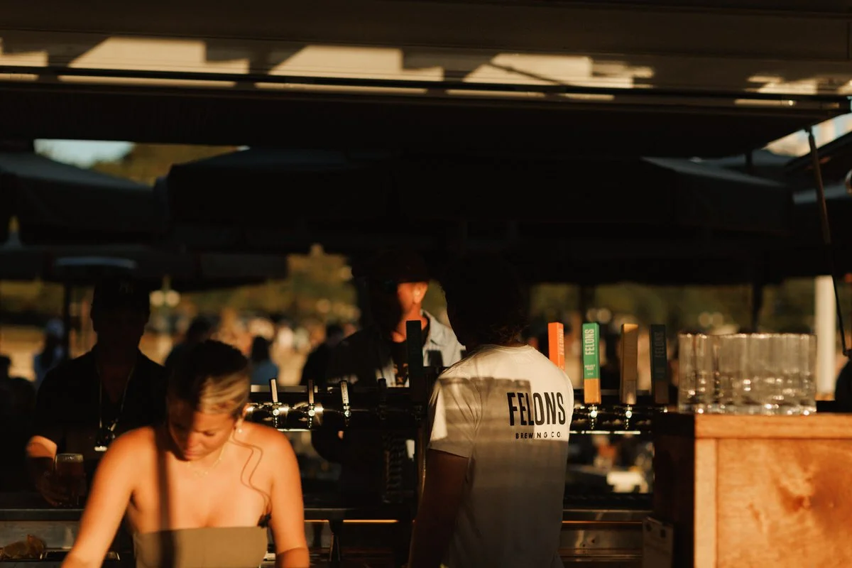 People working at an outdoor bar or brewery stall with beer taps, glasses, and a Felons Brewing Co. t-shirt, under a canopy during sunset.