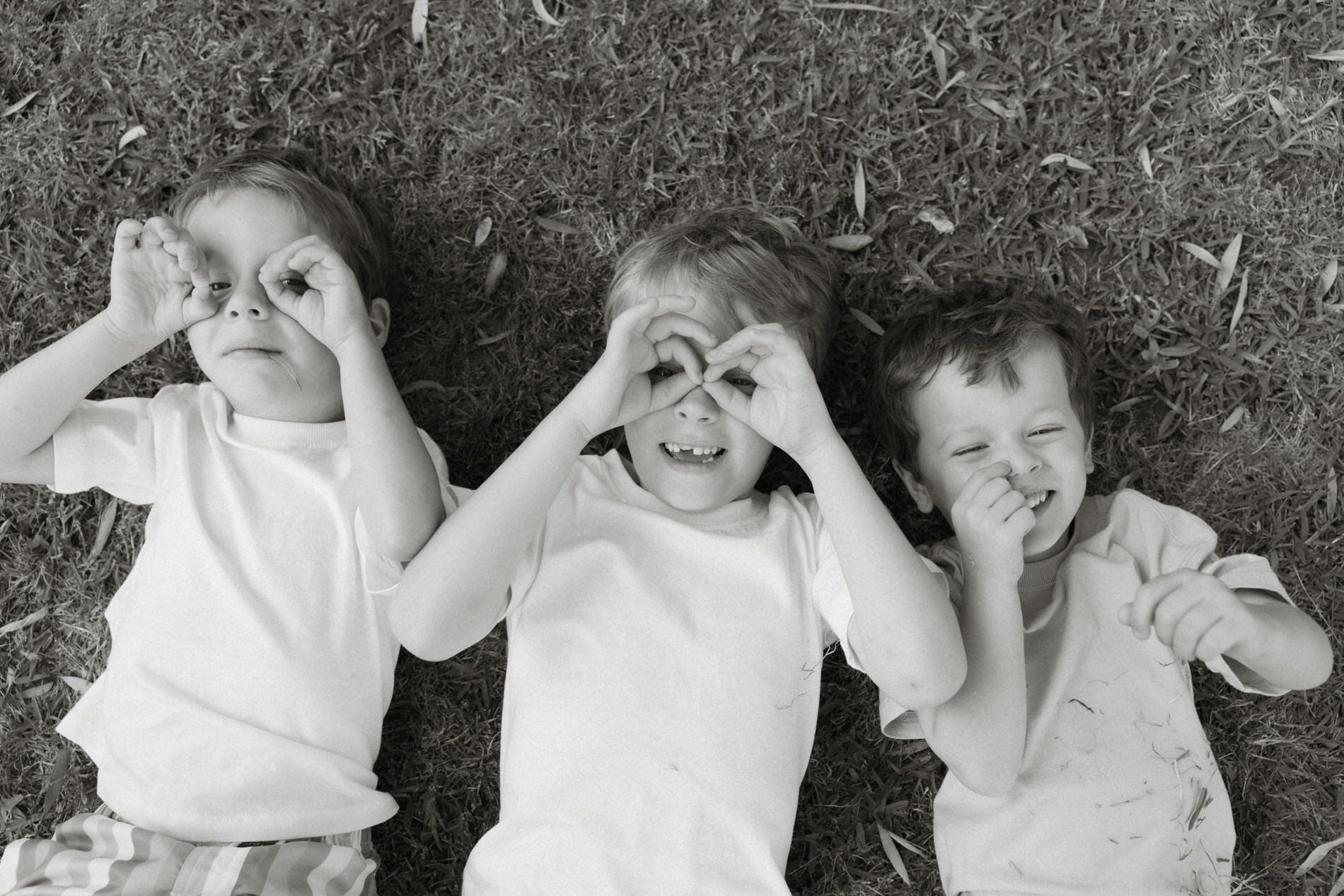 Three children lying on grass making heart shapes with their hands over their eyes, smiling and having fun.