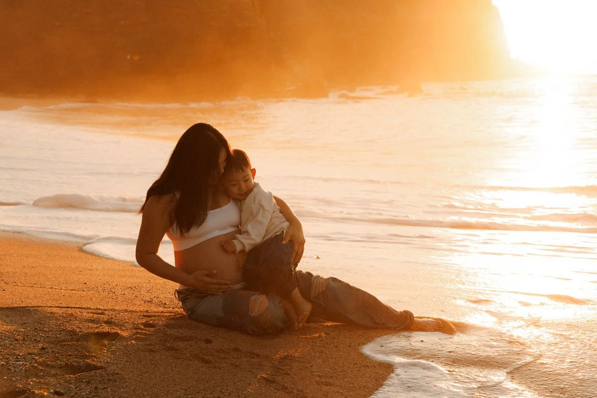 A woman and a young boy sitting on a sandy beach at sunset, with the woman touching her pregnant belly and the boy resting against her.