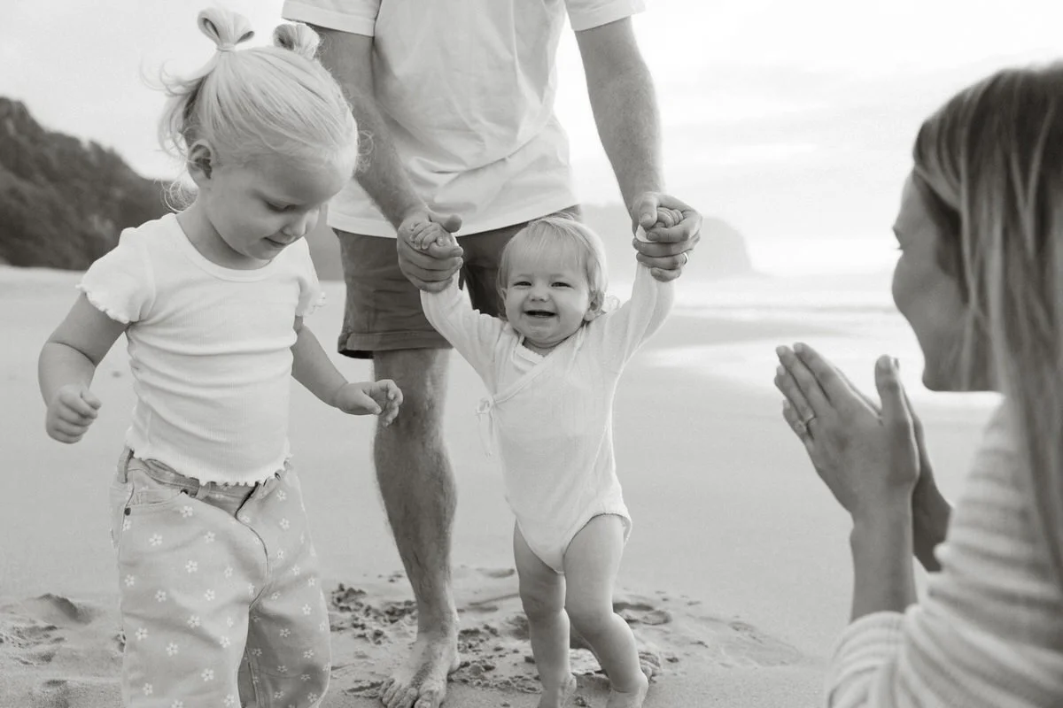 A family on the beach: a man is holding a young child's hands, helping her walk, while a girl and a woman watch and smile.