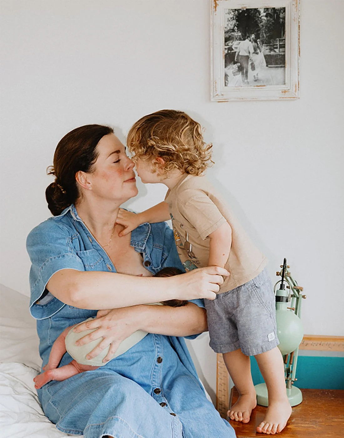A woman and a young boy sharing a tender moment, with their noses touching, inside a home with a framed photo on the wall and a green lamp on a wooden table.