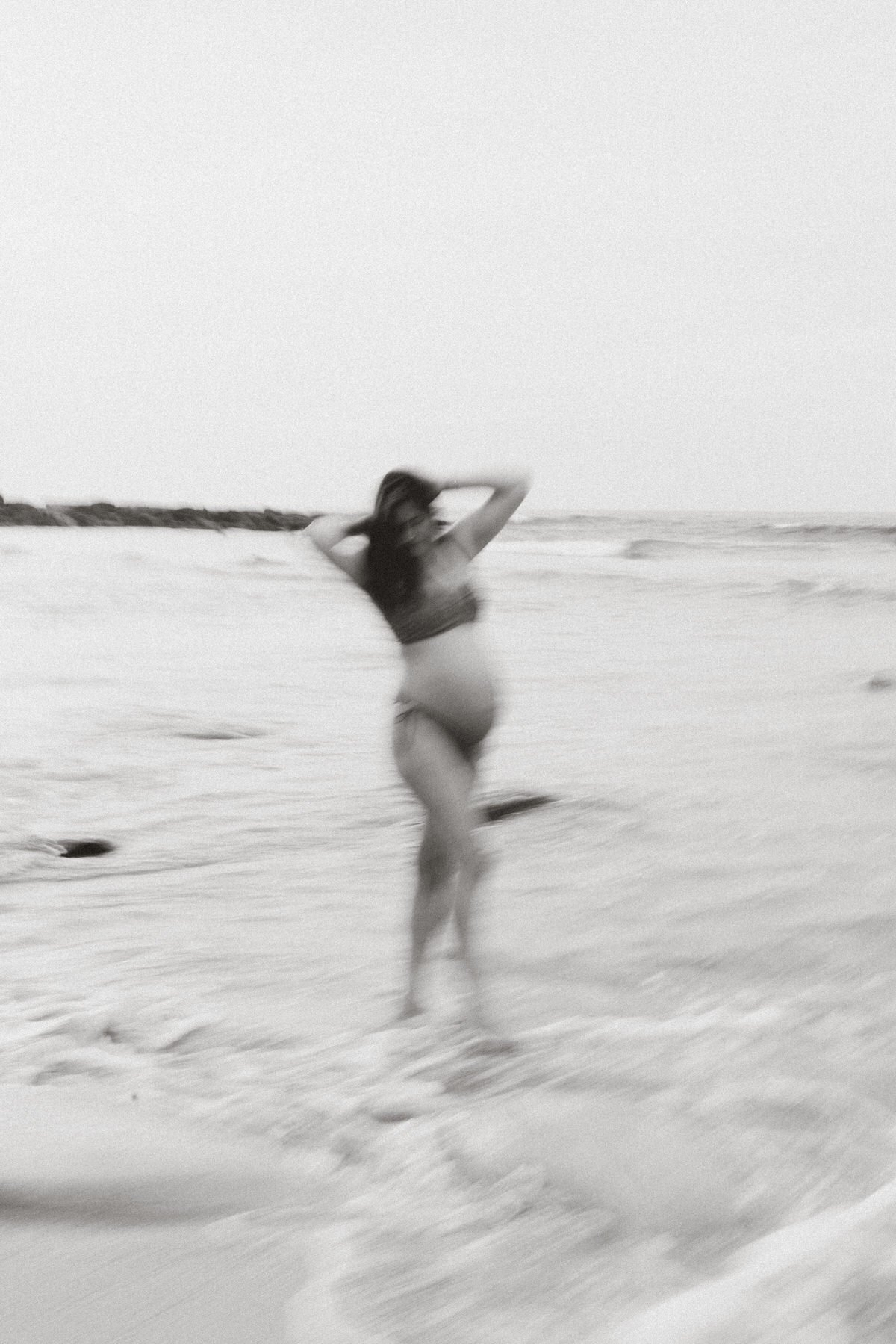 A woman in a swimsuit standing on a sandy beach with ocean waves in the background, blurred motion.