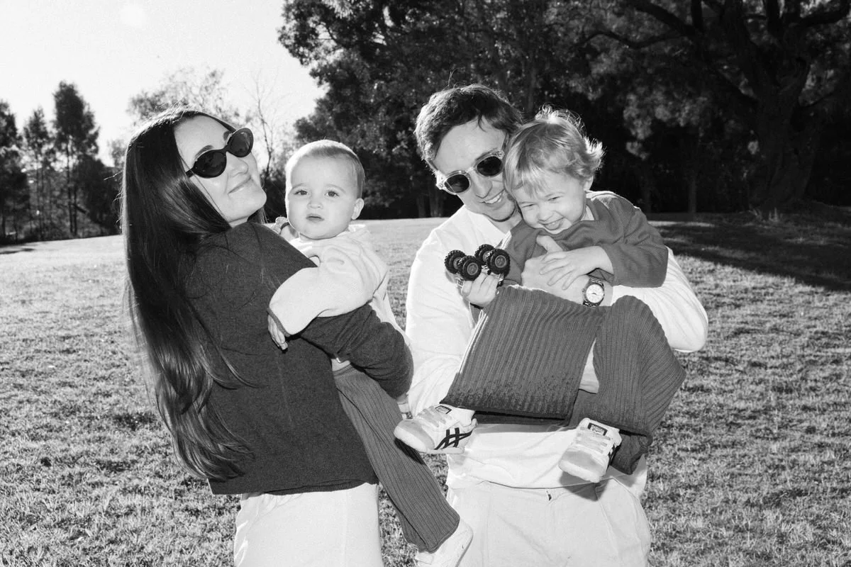 A family of four outdoors in a park, two adults and two young children, all smiling and grabbing each other, with trees in the background, in black and white.