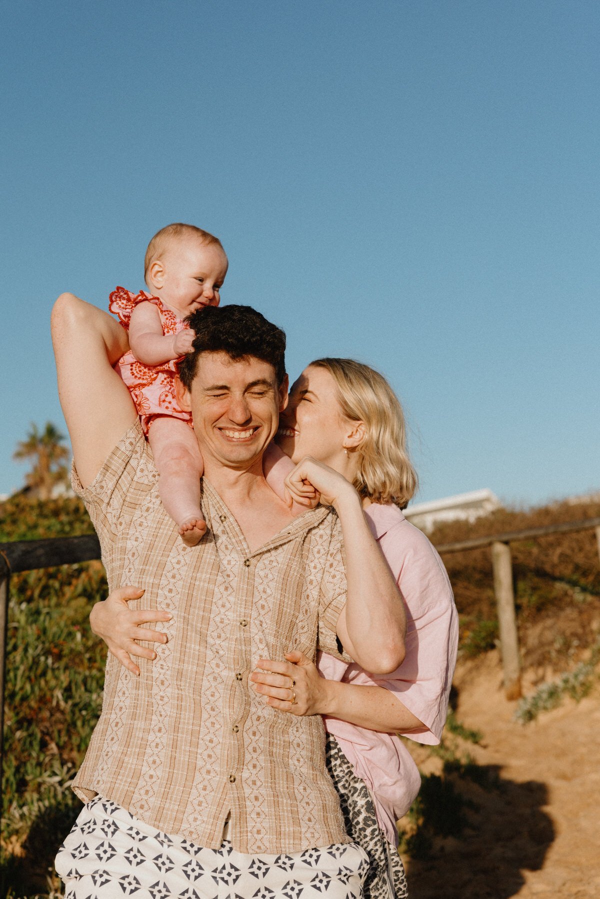A happy family of three enjoying outdoors on a sunny day: a man carrying a baby on his shoulders, a woman kissing the man’s cheek, all smiling in front of a blue sky and greenery.