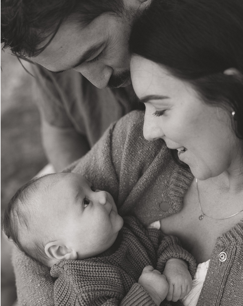 A black-and-white photo of a young family featuring a baby, mother, and father. The mother is holding the baby, and all three are smiling and looking at each other.