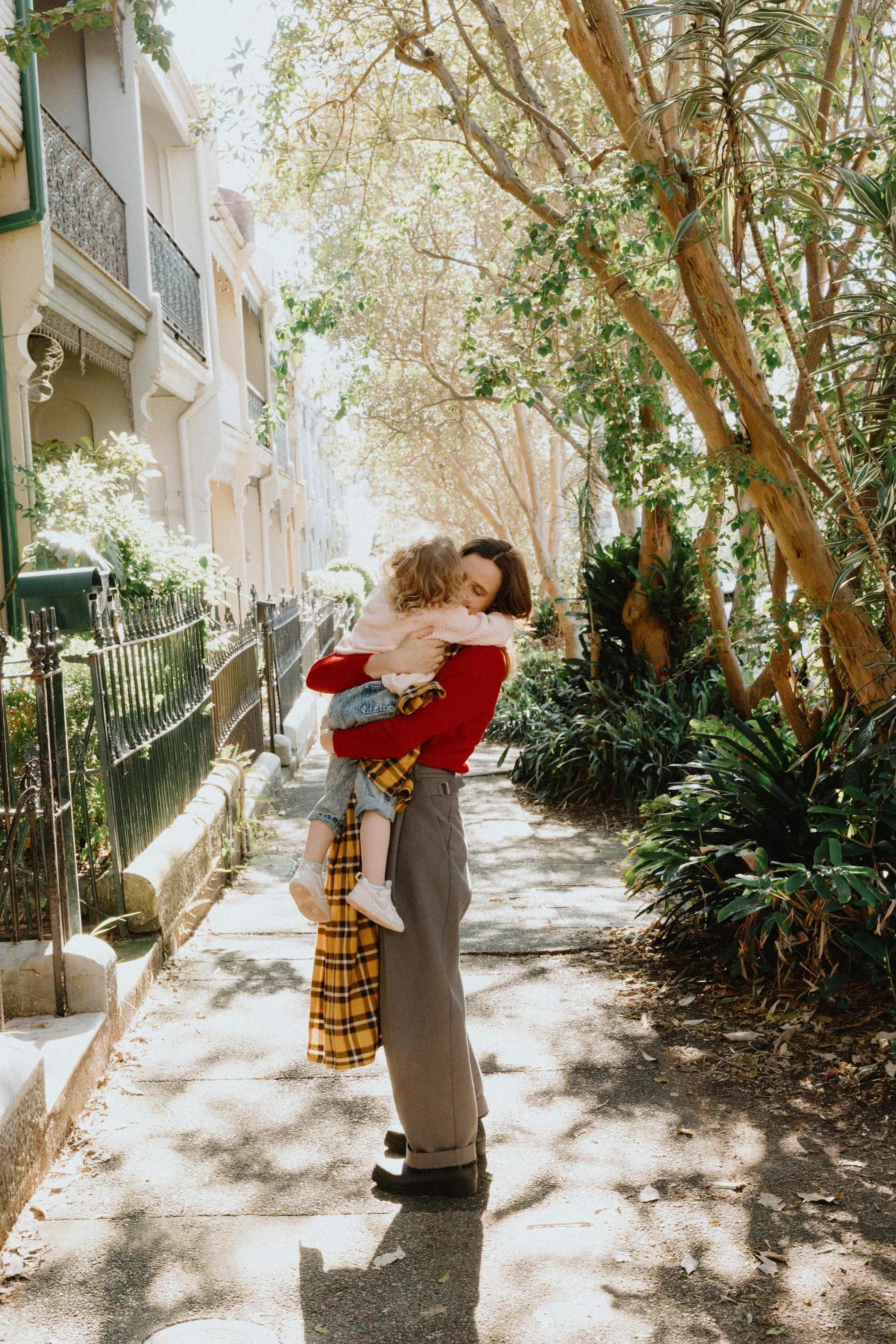 A woman with dark hair and a woman with blonde hair embracing on a sidewalk in a residential area with trees and old buildings, sunlight filtering through the leaves.
