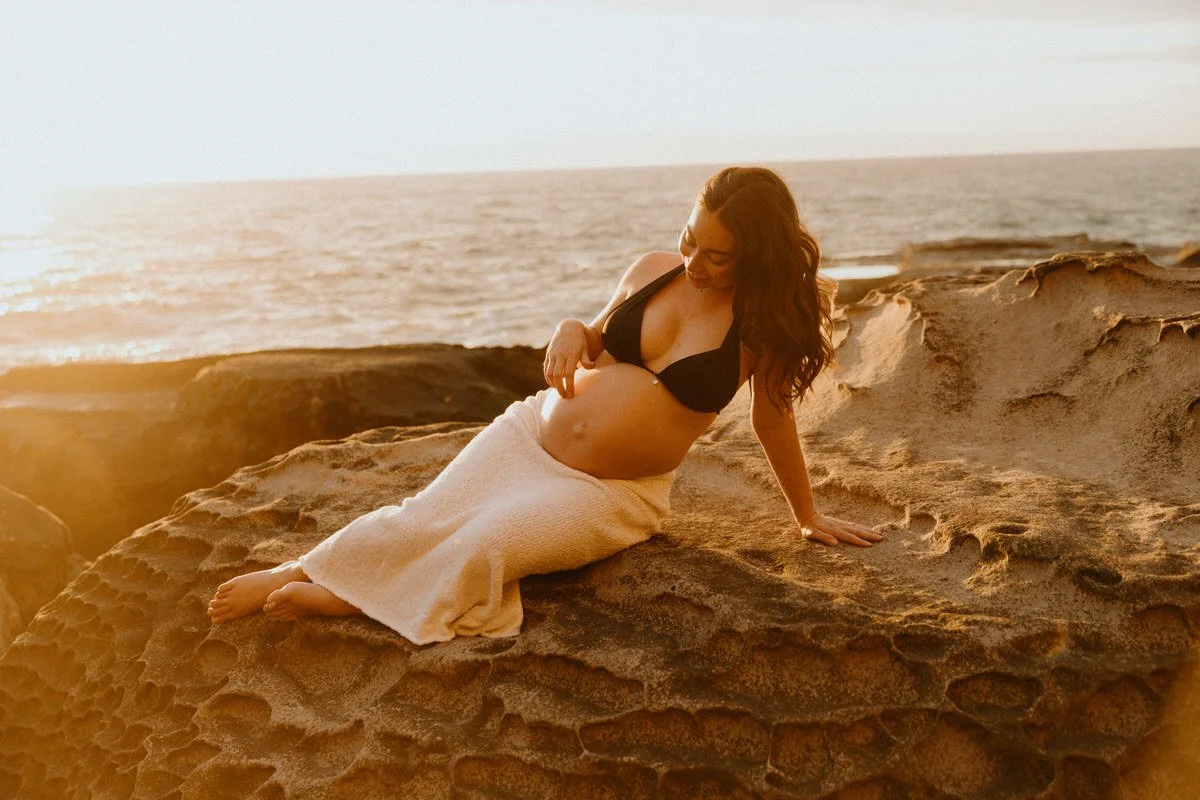 A pregnant woman with long, wavy hair is sitting on a large rock by the ocean at sunset, wearing a black bikini top and a beige skirt.