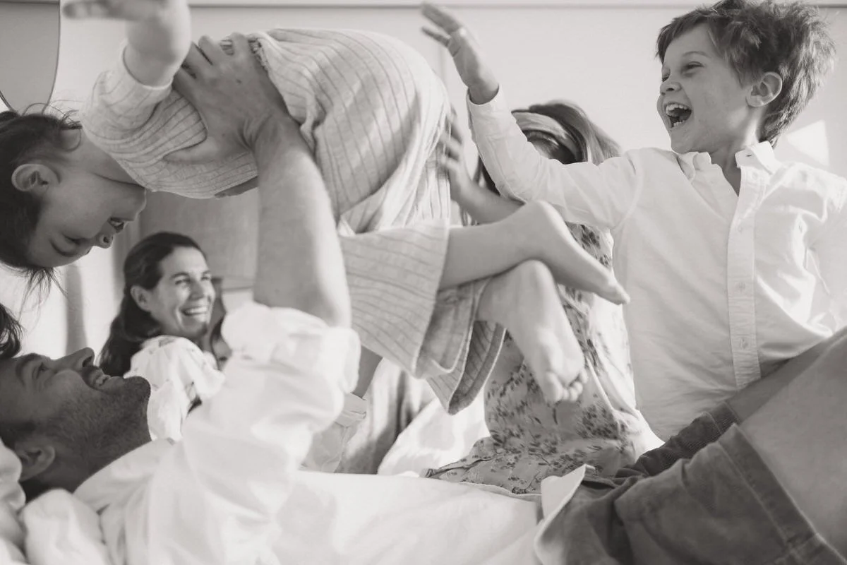 Family playing and laughing together in a hospital room, with children and adults engaging in a playful wrestling or pillow fight.