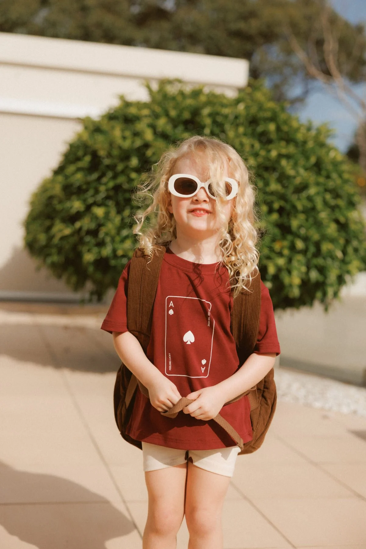 A young girl with curly blonde hair wearing oversized white sunglasses, a red t-shirt with a playing card design, beige shorts, and a brown backpack standing outdoors in front of a large green bush.