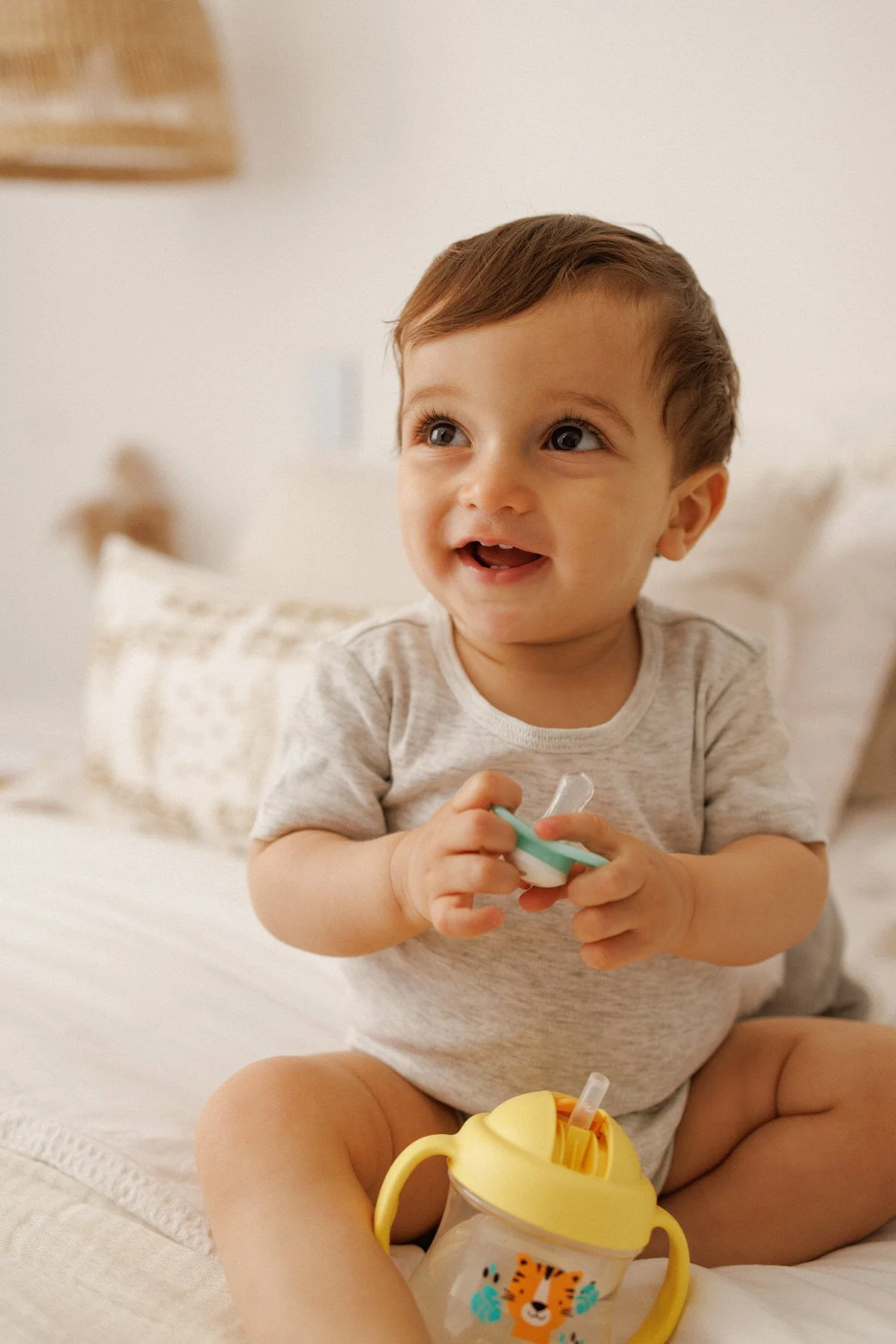 A young boy sitting on a bed, smiling and holding a pacifier with a straw, with a yellow sippy cup featuring a tiger face design in front of him.
