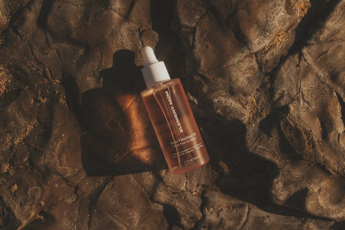 A bottle of Elemnt Mom Belly Fabulous Oil on a rocky surface, casting a shadow.