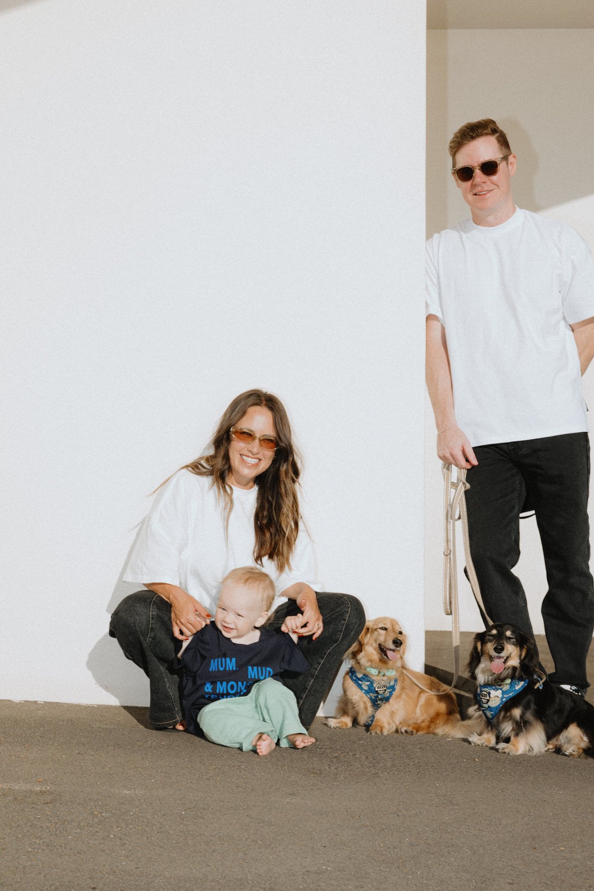 Woman with long brown hair wearing sunglasses, a white T-shirt, and black jeans, sitting on the floor with a young girl, a toddler, and three dogs. The woman and the girl are smiling, and the toddler is looking away with a curious expression. The man