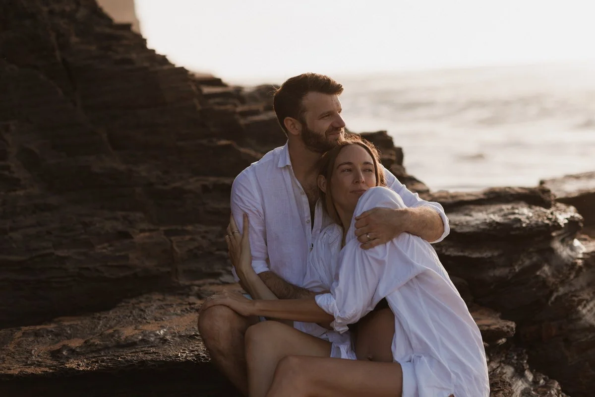 A happy couple sitting on rocks by the ocean during sunset, embracing each other peacefully.
