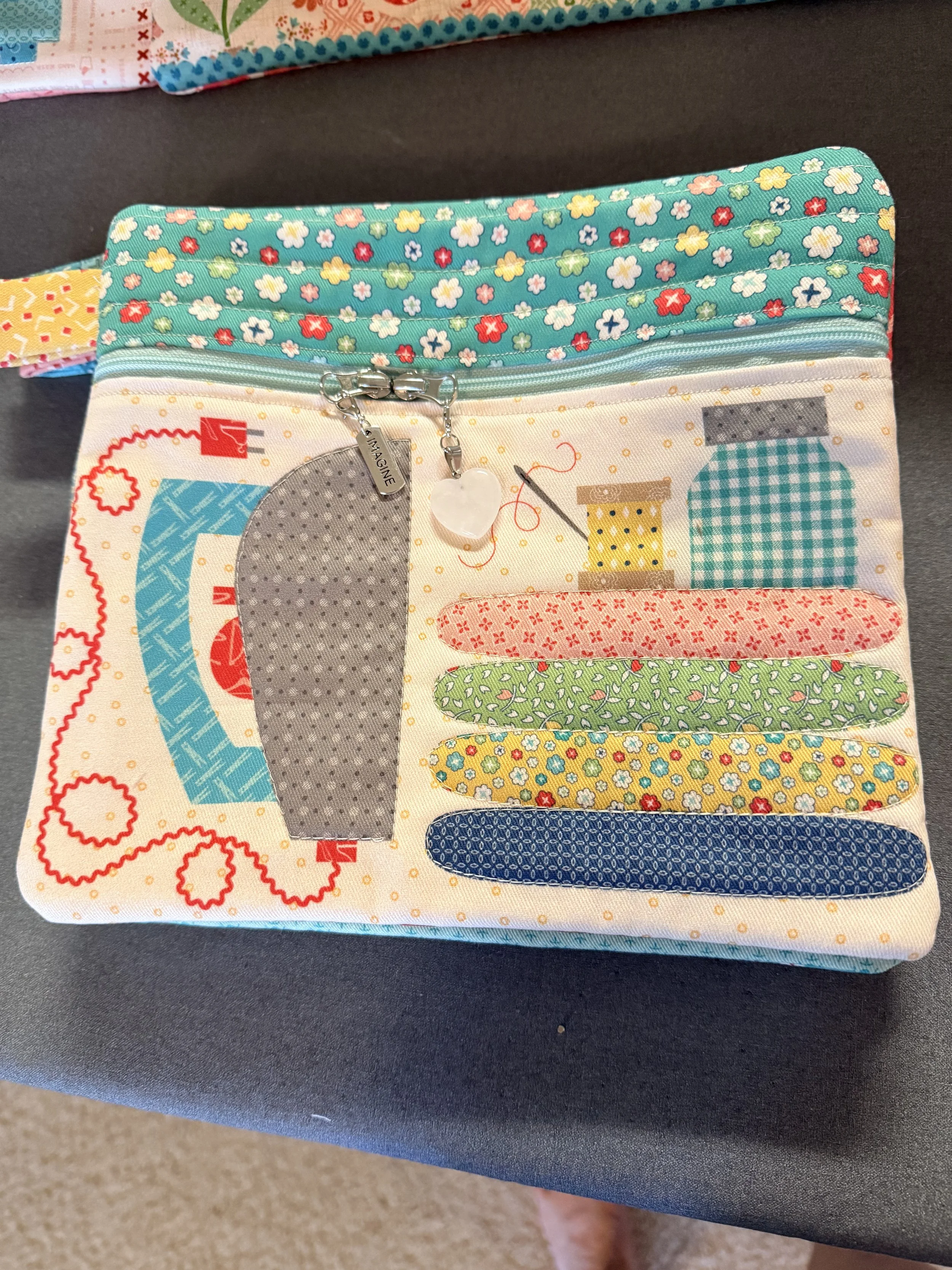 A fabric organizer with multiple colorful compartments featuring various fabric patterns and a silver zipper with a heart-shaped pull, placed on a dark surface.
