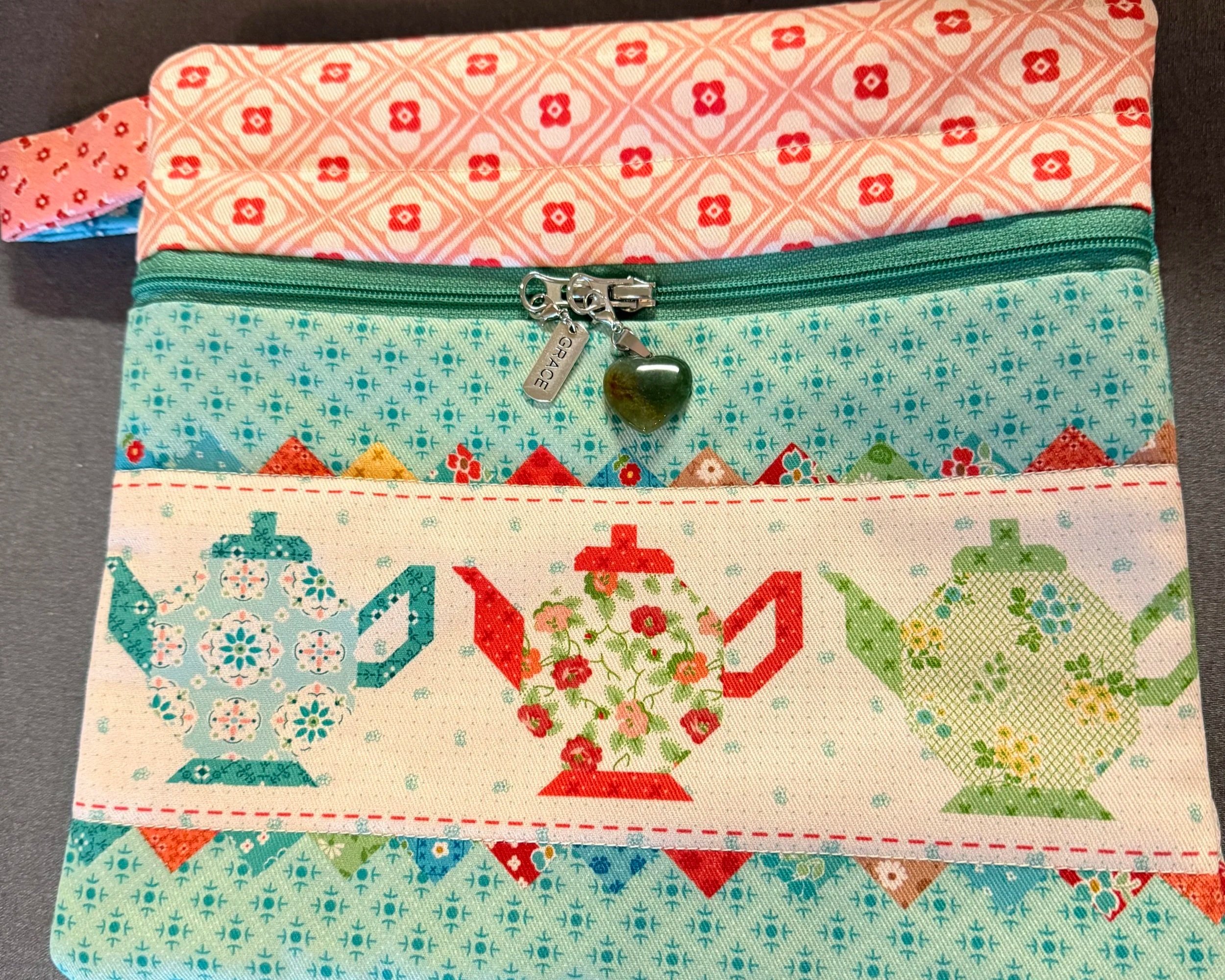 A fabric pouch with a floral and geometric print, a green zipper with a heart-shaped pull, and a decorative chain with a tag that says 'GRACE'.