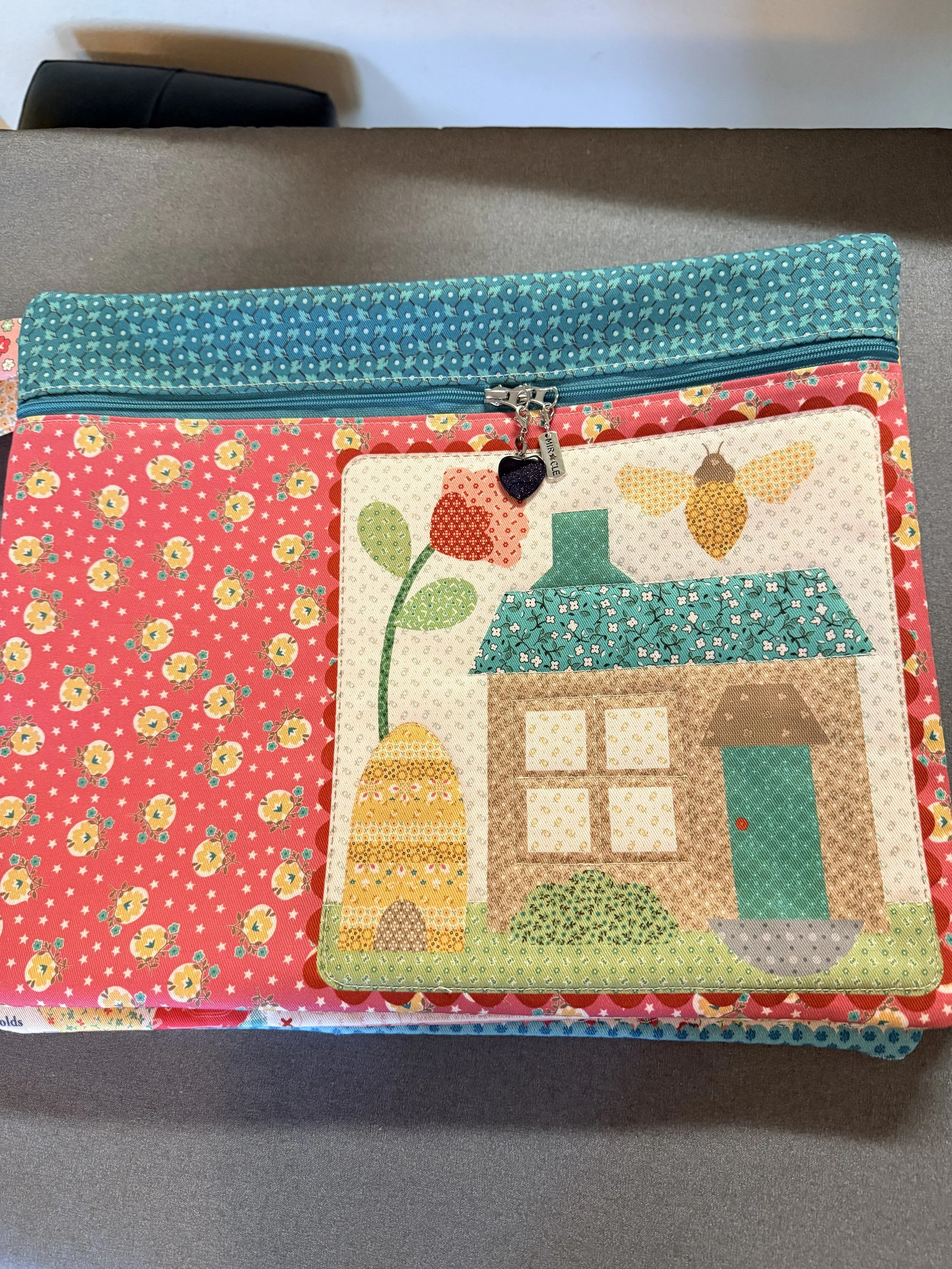 Colorful fabric pouch with a zipper on top, decorated with a patch featuring house, bee, and flower motifs, placed on a gray surface.