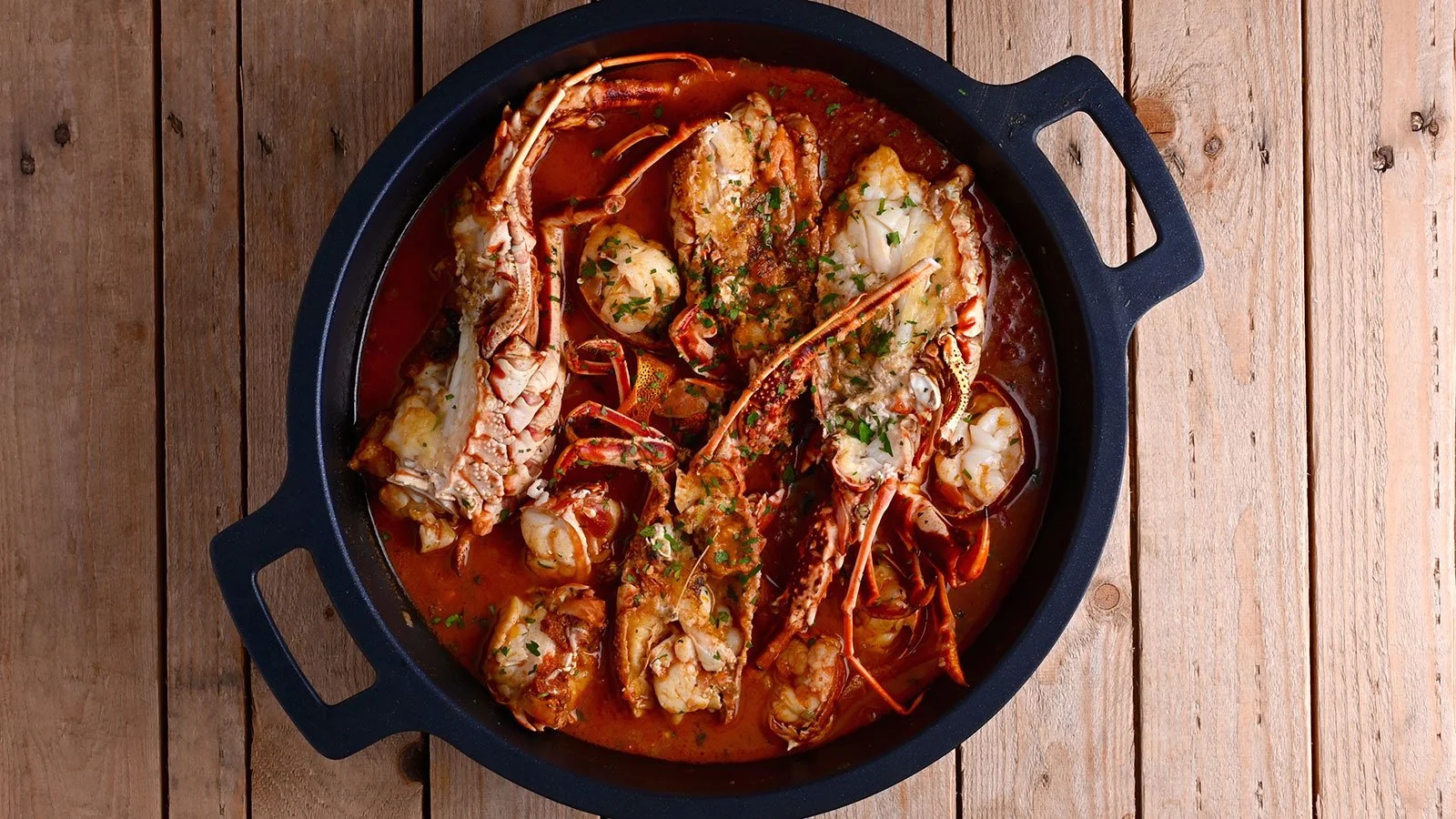 Seafood boil in a black skillet on a wooden surface, including lobster, shrimp, crab, and potatoes in a red cocktail sauce