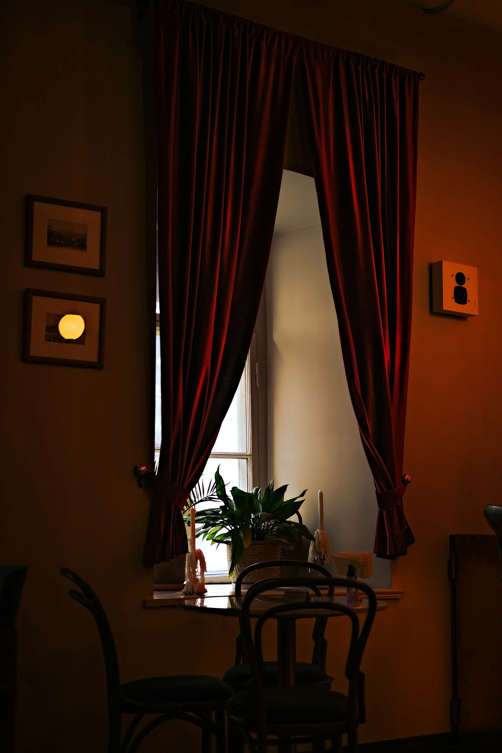 Une pièce éclairée par la lumière naturelle d'une fenêtre encadrée de rideaux rouges. Sur la fenêtre, une plante verte sur la table, avec deux bougies et un cadre photo. Plusieurs chaises entourent la table.