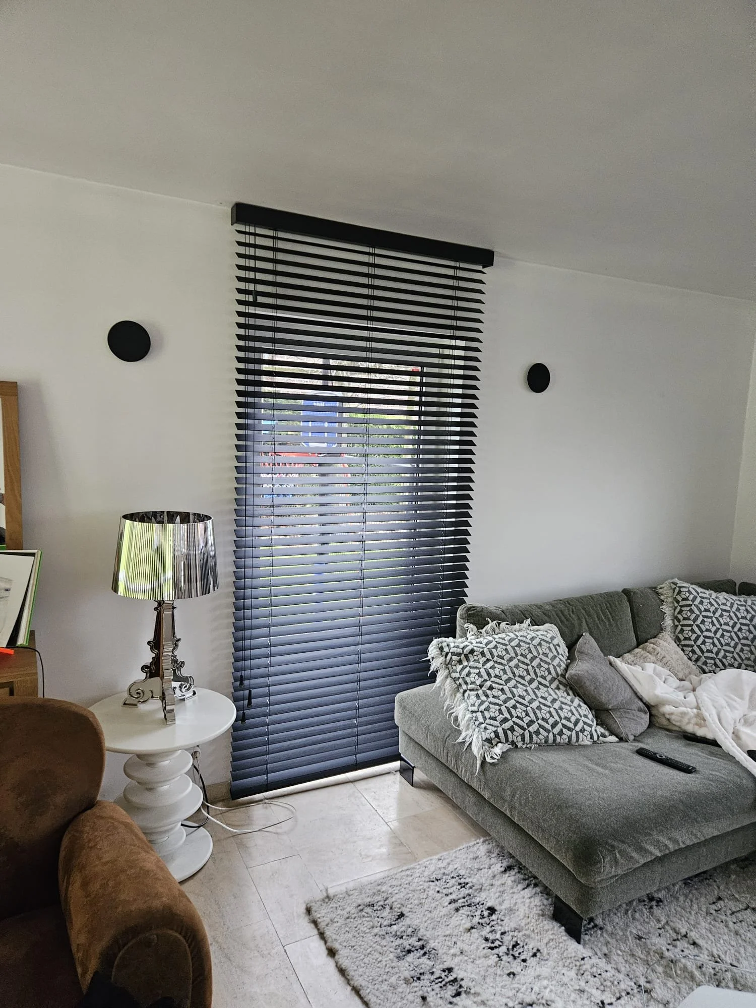 Salon moderne avec un canapé gris, des coussins géométriques, un tapis en laine blanche, une lampe avec abat-jour en métal et des stores à rayures noires et blanches devant une fenêtre.